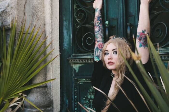 A-woman-with-long-blonde-hair-and-tattooed-arms-raises-her-hands-overhead-against-a-weathered-teal-door-framed-by-palm-fronds Best Trash Polka Tattoo Artists in NYC (2026)